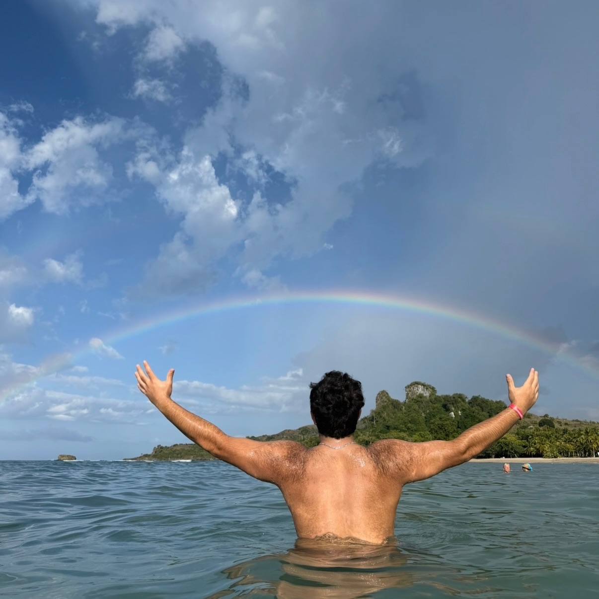 Antony from behind, arms wide, standing in the sea beneath a rainbow.