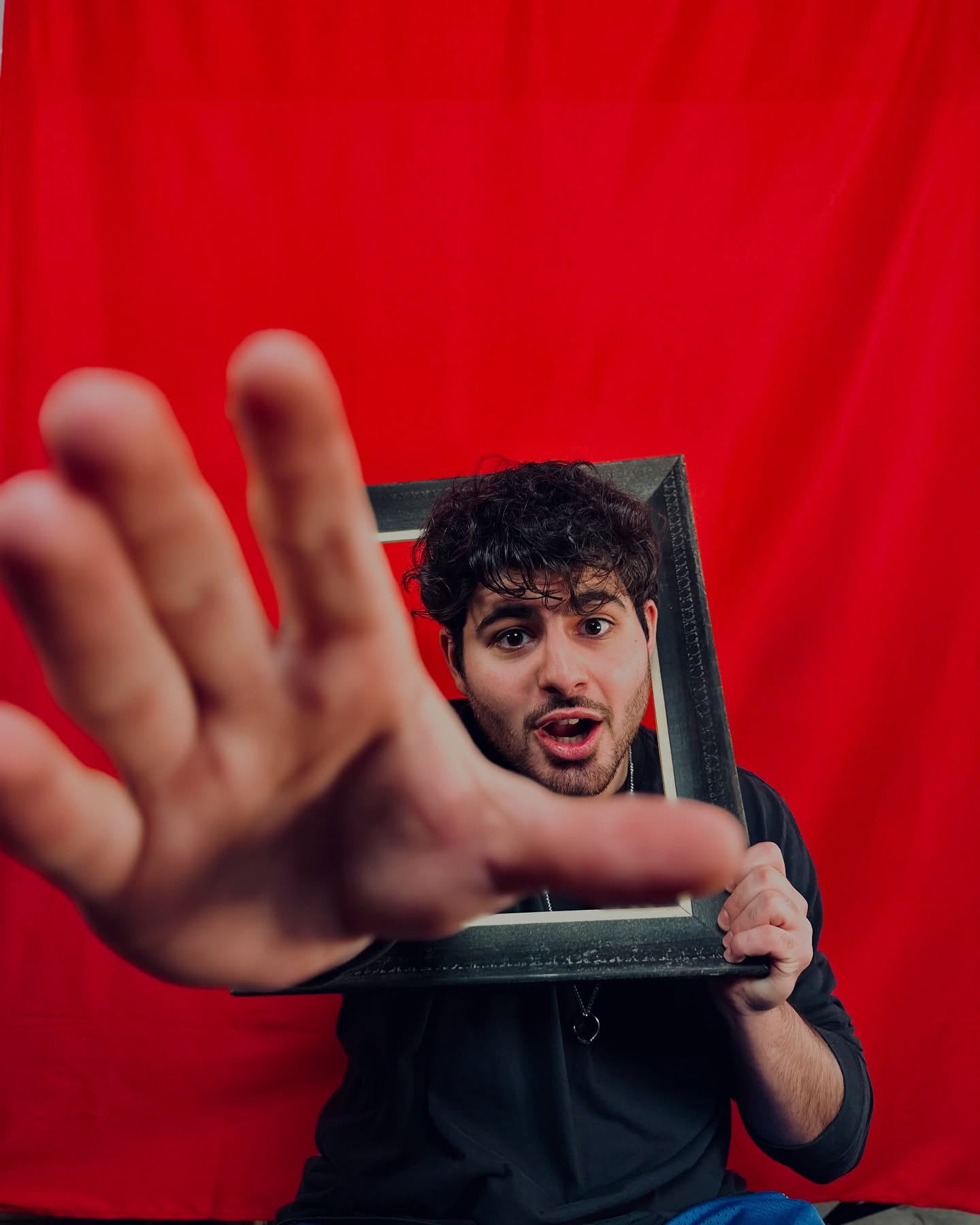 Antony holding an empty picture frame against a red backdrop, one hand reaching toward the camera.